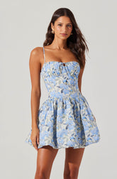 A woman is wearing a sleeveless, fitted dress with thin spaghetti straps. The dress features a square neckline with a small keyhole cutout at the center, and the fabric has a light blue and white floral pattern with hints of darker blue. The waistline is slightly cinched, and the skirt is voluminous and gathered, ending above the knee.